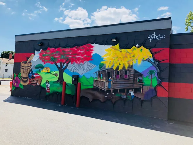 The mural on El Pilón Criollo's wall: the black cinderblock cracks open onto a Puerto Rican countryside — a pilón, a red flamboyán tree, a jíbaro's casita and blue mountains